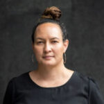 Portrait of Fiona Cornforth, a woman with a calm expression, wearing a black top and large hoop earrings. She has dark hair styled in a bun and stands against a plain, dark background.