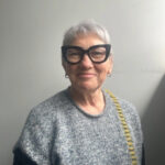 Portrait of Juanita Sherwood, an older woman with short grey hair and wearing large, thick black-framed glasses. She is dressed in a grey knit sweater with a patterned strap over her shoulder, standing against a neutral background.