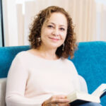 Portrait of Dr. Nicole Tujague, a woman with curly brown hair, smiling while sitting on a blue chair. She is wearing a light-colored top and holding an open book in her hands, with a soft, neutral background behind her.