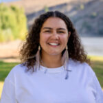 Portrait of Tanika Perry, a young woman with curly brown hair, smiling outdoors. She is wearing a white top and long feather earrings, with a natural background featuring trees and hills.