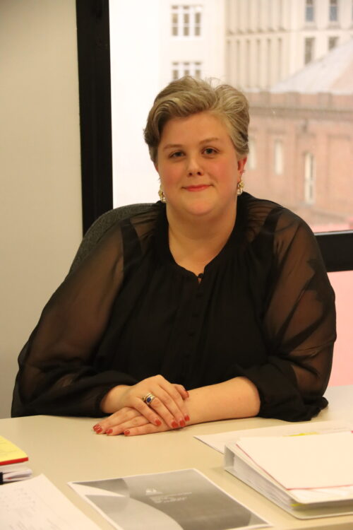 A person with short, grey-blonde hair wearing a sheer black blouse sits at a desk with hands folded. They are smiling softly, wearing gold earrings and a blue ring. Papers and documents are spread out on the desk, and a window behind them shows tall city buildings.