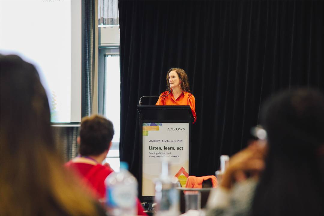 Dr Tessa Boyd-Caine, CEO of ANROWS, stands at a podium speaking during the ANROWS Conference 2025. The podium displays the conference theme “Listen, learn, act.” Audience members are visible in the foreground, slightly out of focus.