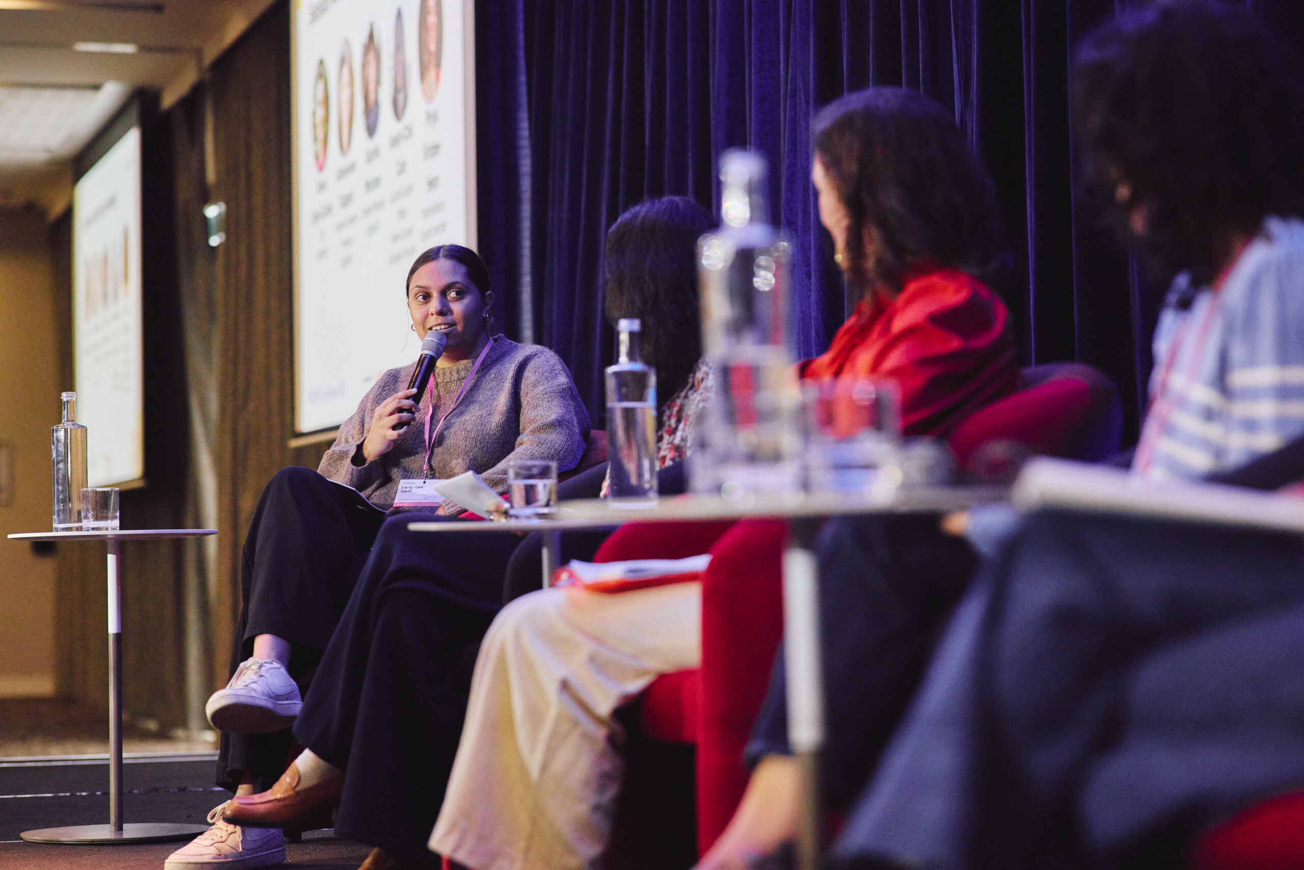 Keely-Chè Cain speaks into a microphone during the Listen for our healing panel on Friday 16 May at the ANROWS Conference 2025. Seated on stage to the left, Keely-Chè wears a grey jumper and dark trousers, with a lanyard visible around her neck. She faces three other panellists, who are partially visible in the foreground and to the right, seated in red chairs. The backdrop is a deep navy curtain, and a projected slide showing panellist names and images is visible in the background. Water bottles and glasses rest on small tables between the speakers.