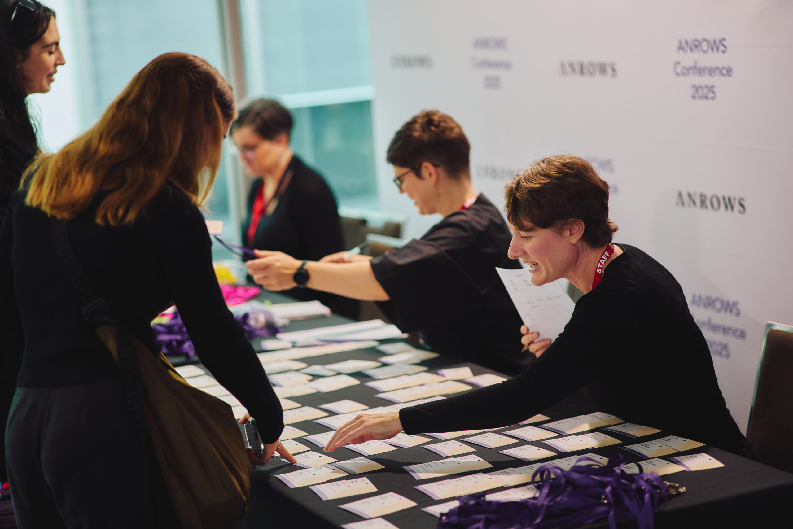 Dr Nicole Weeks, Research Manager for NCAS at ANROWS, is warmly welcoming attendees at the registration desk on Friday 16 May during the ANROWS Conference 2025. Positioned in the foreground on the right, Nicole smiles and gestures toward a name badge on the table while holding a registration list. Rows of name tags and purple lanyards are spread across the table. Behind her, other ANROWS staff assist additional attendees. The backdrop features a branded ANROWS Conference 2025 media wall. The scene is bright and welcoming, capturing the energy of the event's opening moments.