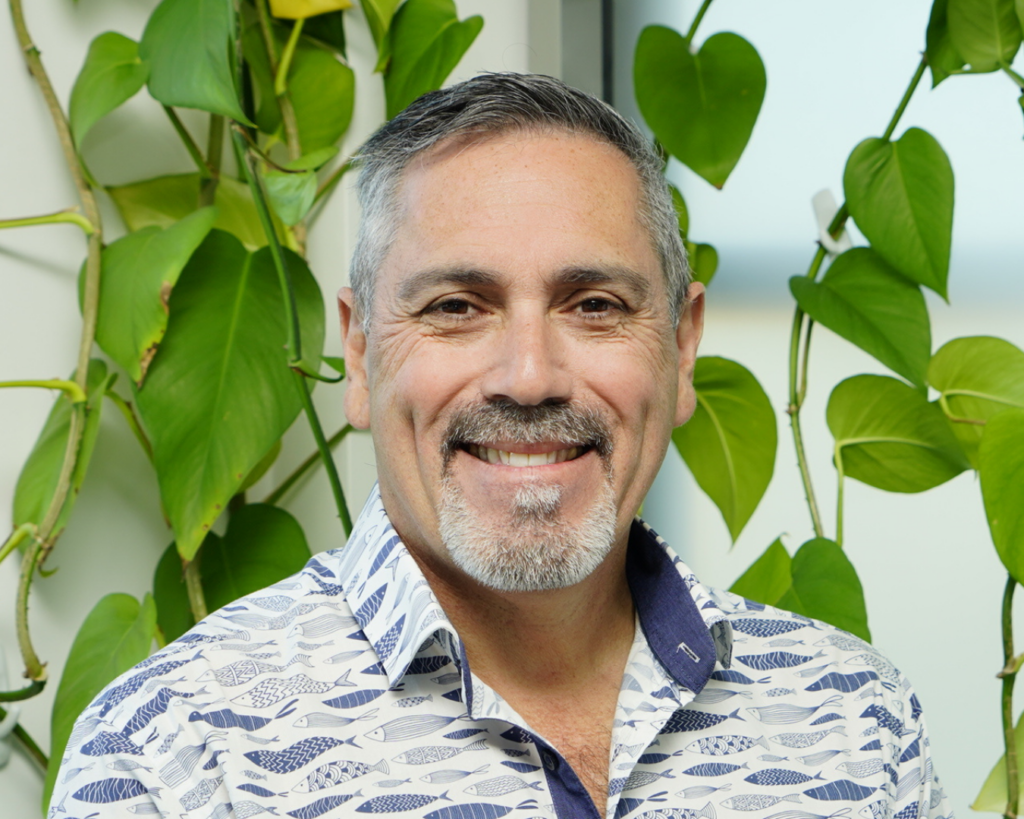 Portrait of Craig, smiling at the camera in front of green vines.